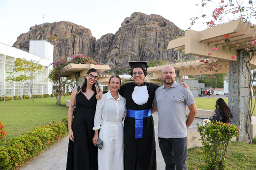 IMagem: um grupo de quatro pessoas posa para uma foto ao ar livre, com uma paisagem de montanhas ao fundo. A pessoa ao centro está vestida com beca e capelo de formatura, simbolizando um momento de conquista. Ela é acompanhada por três familiares: uma mulher de vestido preto, uma mulher de roupa branca e um homem de camiseta cinza. Todos estão sorrindo, transmitindo felicidade e orgulho.