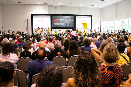 Image: Foto mostra o público de costas no Centro de Convivência do Campus do Pici lotado; no palco, apresenta-se o grupo Maracatu Solar, em frente a um telão. (Foto: Álvaro Graça Jr./ UFC Informa)