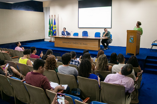 Imagem: Foto com a Profª Zelma Madeira em pé no púlpito fazendo uso da palavra, enquanto o público assiste à aula magna