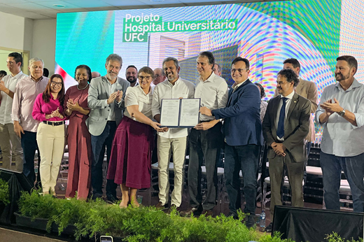 Imagem: De pé, no palco, ladeados por outras autoridades, estão Arthur Chioro (presidente da EBSERH), Josenília Gomes (superintendente do Complexo Hospitalar), Elmano de Freitas (governador), Camilo Santana (ministro da Educação) e Custódio Almeida (reitor da UFC). Eles seguram a ordem de serviço e o termo de autorização de licitação assinados