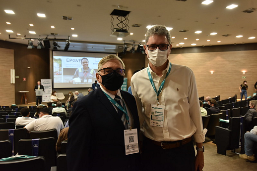 Imagem mostra os professores Fernando Nunes e Augusto Albuquerque em pé e lado a lado no auditório, com telão e pessoas sentadas ao fundo