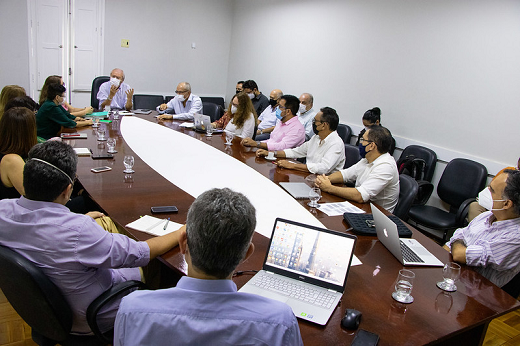 Imagem mostra várias pessoas sentadas em volta de uma mesa em formato oval, com bordas de cor marrom e centro de cor branca