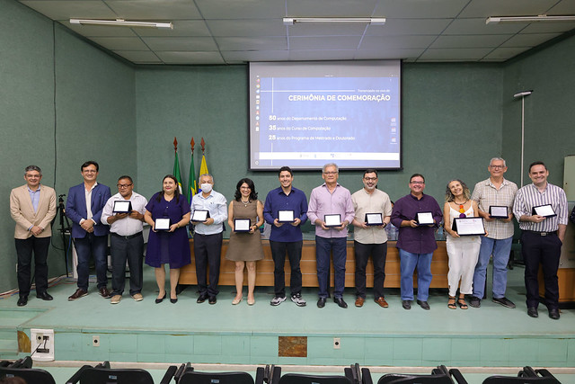 imagem: Grupo de pessoas posa para foto no auditório do Centro de Ciências da Universidade Federal do Ceará. Elas estão lado a lado, sorrindo, e seguram placas de homenagem. Ao fundo, há um telão com o título “Cerimônia de Comemoração: 50 anos do Departamento de Computação, 35 anos do Curso de Computação e 25 anos do Programa de Mestrado e Doutorado”, além das bandeiras do Brasil, do Ceará e da Universidade.