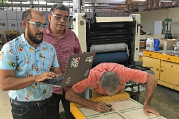 Imagem: foto do prof Chico Neto segurando um notebook e observando os diplomas sendo impressos