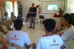 Imagem: Foto de aula do curso com professor de pé e estudantes sentados em carteiras em círculo, ao fundo, tela com projeção