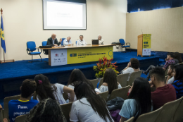 Imagem: Foto do auditório com parte do público e palestrantes sentados à mesa do evento