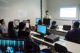 Imagem: Foto de servidores assiatindo a uma aula num laboratóriode informática. Podemos vê-los sentados e, frente a msas com computadores e, à frente de todos, o professor apresentando conteúdo em power point no quadro. 