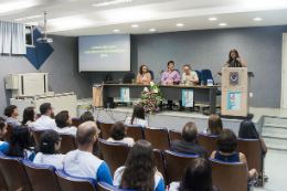 Imagem: Foto a partir do fundo do auditório José Albano, onde se vê o público sentado e a mesa no palco com professores