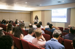 Professora em pé palestra para plateia, frente a uma apresentação de PowerPoint. 