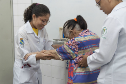 Duas integrantes do Hospital Universitário ajudando uma idosa a se locomover em uma sala do hospital (Foto: Viktor Braga/UFC)