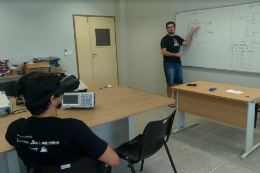 Imagem: foto de um estudante sentado com óculos observando o professor dando explicações na lousa dentro de uma sala de aula