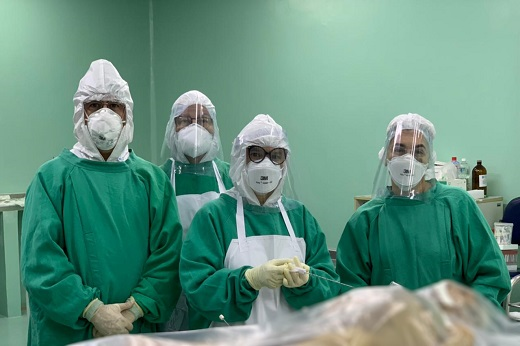 Equipe que realizou a autópsia, totalizando quatro pessoas, posicionadas lado a lado; elas estão em uma sala e vestem uniformes verdes, máscaras e toucas; em primeiro plano, há um cadáver coberto com saco plástico, sendo que imagem do cadáver foi desfocada (Foto: Divulgação)