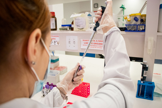 Imagem: foto e uma jovem de cabelos presos e de máscara descartável segurando um material de laboratório