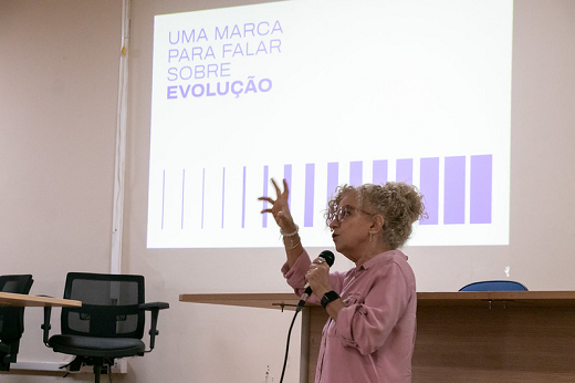 imagem mostra a professora Cláudia Buhamra na parte inferior fazendo a apresentação, enquanto ao fundo, na parte superior, aparece uma projeção na parede 