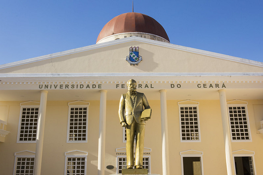 Imagem mostra parte da fachada do campus de Sobral da UFC, com estátua na cor dourada a frente e céu azul ao fundo