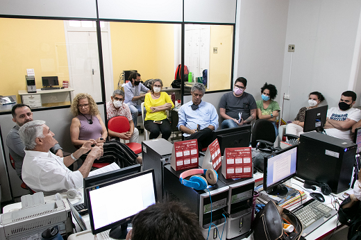 Imagem: Cerca de dez pessoas em uma sala da Rádio Universitária, sentadas em cadeiras dispostas em formato de semicírculo; ao lado há móveis com computadores e outros equipamentos de trabalho da equipe da emissora (Foto: Viktor Braga/UFC)