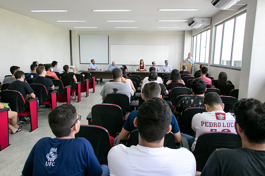 Imagem: Pessoas sentadas em cadeiras no auditório, com uma mesa de autoridades adiante