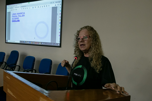 Imagens: A Profª Cláudia Buhamra falando ao microfone, em pé, no auditório da Reitoria; no fundo há uma tela com a marca do PDI (Foto: Viktor Braga/UFC)