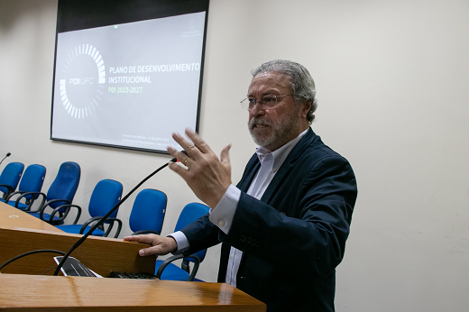 Imagem: Reitor em pé, falando ao microfone no auditório da Reitoria; ao fundo há uma tela com a projeção da marca do PDI (Foto: Viktor Braga/UFC)