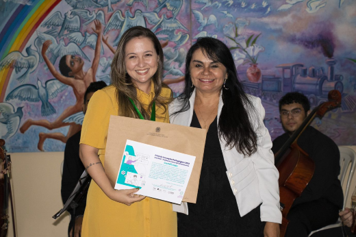 Imagem: Foto da Professora Carolina Morais, da Casa de Cultura Britânica da UFC, recebendo da coordenadora do PAAP, Mazzé Santos, o primeiro lugar do Prêmio INOVPED.