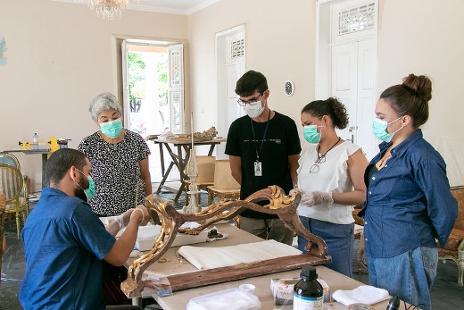 Imagem: na foto, cinco pessoas estão em um salão observando as pernas de uma cadeira dourada que está sobre uma mesa. Todos usam máscaras e um jovem sentado limpa o objeto com um pincel