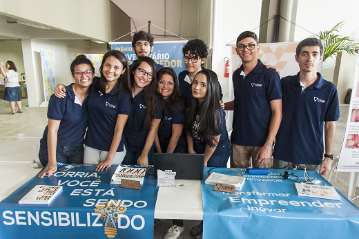 Imagem: Grupo de nove estudantes, em pé, atrás de uma mesa na qual estão expostas informações sobre um trabalho realizado pela equipe (Foto: Viktor Braga/UFC)