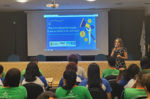 Imagem: foto de jovens de blusa verde estão de costas, observando uma mulher loira, jovem apresentar um trabalho em frente ao data show