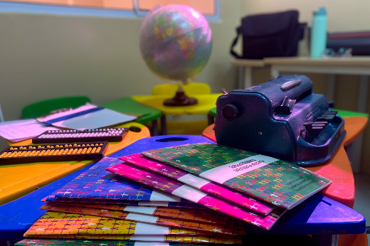 Imagem: na foto, cartilhas estão dispostas sobre uma mesa azul. Ao lado, uma máquina de datilografar braille e, ao fundo, um globo terrestre compõem o cenário