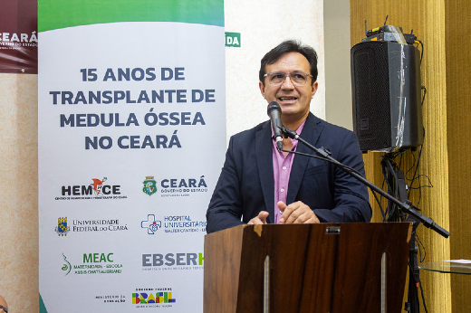 Imagem: Reitor Custódio Almeida discursando no púlpito do auditório, e ao fundo um banner com os dizeres "15 anos de transplante de medula óssea no Ceará"