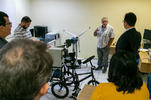 Imagem: Um grupo de pesquisadores em pé, sendo apresentados a um protótipo de um triciclo assistivo para crianças com hemiplegia