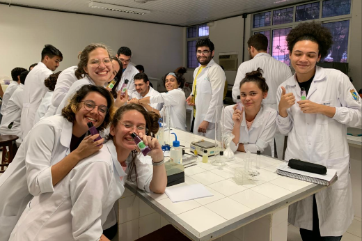 Imagem: A imagem mostra um grupo de estudantes em um ambiente de laboratório, todos vestindo jalecos brancos, sugerindo que estão em uma aula prática de ciências ou biologia. A maioria das pessoas está sorrindo, e algumas seguram tubos de ensaio coloridos, aparentemente realizando algum experimento ou demonstrando satisfação com a atividade.  O ambiente é iluminado, e as bancadas possuem equipamentos de laboratório, como pipetas e frascos. Ao fundo, há mais estudantes envolvidos na atividade. A atmosfera parece descontraída e colaborativa, com os alunos interagindo e se divertindo durante a experiência.