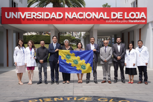 Imagem: A imagem mostra um grupo de pessoas em frente à entrada da Universidad Nacional de Loja. O nome da universidade está destacado em um letreiro vermelho acima do grupo, junto com o slogan "Educamos para Transformar". O grupo é composto por indivíduos vestidos tanto com trajes formais quanto com jalecos médicos, sugerindo que são profissionais ou membros da equipe acadêmica. Eles estão segurando uma bandeira azul e amarela com um design de emblemas verdes ou estrelas no centro. O cenário parece ser uma área externa da universidade, com um logotipo ou emblema visível no chão à frente deles, além de palmeiras e a entrada de um prédio ao fundo.