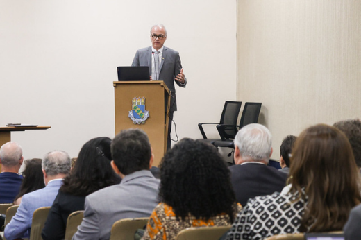 Imagem: Foto do reitor Daniel Diniz, da UFRN, discursando no púlpito do Auditório da Reitoria, vestindo um paletó cinza