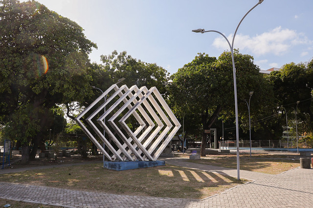 Imagem: foto de uma praça com um símbolo de quatro losangos brancos. Atrás, há árvores e, ao lado, um poste de luz