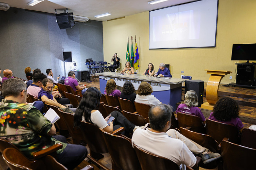 Imgem: A imagem mostra o anfiteatro da Faculdade de Direito da UFC, onde ocorreu o seminário. Há uma mesa no palco com quatro pessoas sentadas, participando de uma discussão. Ao fundo, uma tela grande exibe uma apresentação, e algumas bandeiras estão posicionadas ao lado. Na plateia, várias pessoas estão sentadas em cadeiras de madeira, algumas usando camisetas roxas. Alguns participantes estão anotando ou ouvindo atentamente a apresentação. O ambiente tem iluminação artificial e um aspecto formal.