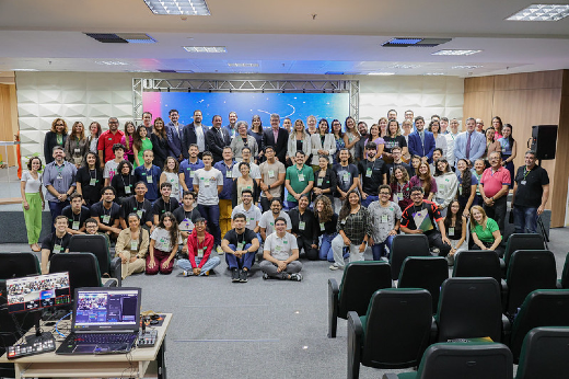 Imagem: A imagem mostra um grande grupo de pessoas reunidas em um auditório para uma foto em grupo: são alunos da UFC, e autoridades da UFC e do TRE/CE. O ambiente possui cadeiras organizadas em fileiras e uma mesa com equipamentos eletrônicos, como notebooks e telas de monitoramento, na frente. No fundo, há um palco com um painel iluminado e uma estrutura metálica. O grupo é diverso, composto por jovens e adultos, muitos usando crachás do hackathon. A maioria das pessoas está sorrindo e posando para a foto.