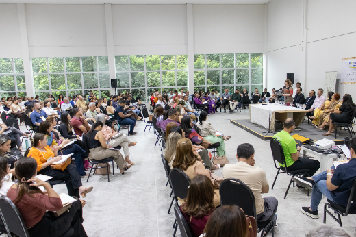 Imagem: foto que exibe uma grande sala com pessoas sentadas em cadeiras e um palco com cerca de 10 pessoas sentadas numa mesa