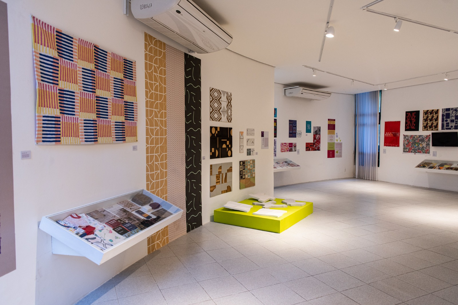 Imagem: A imagem mostra uma galeria de arte (Museu de Arte da UFC) com paredes brancas e piso claro. Diversas obras gráficas e têxteis coloridas estão expostas nas paredes, incluindo tapeçarias, cartazes e padrões geométricos. Vitrines inclinadas exibem materiais gráficos, e no centro há uma base verde com livros abertos. O ambiente é bem iluminado e organizado, com foco em design e arte visual.