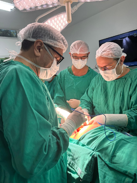 Imagem: foto mostra três médicos em um centro cirúrgico com luz clara e uma pessoa deitada em maca hospitalar com o útero exposto