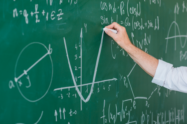 Imagem: Foto de um quadro verde com cálculos matemáticos escritos em giz branco e a mão de uma pessoa branca segurando um giz e escrevendo na lousa