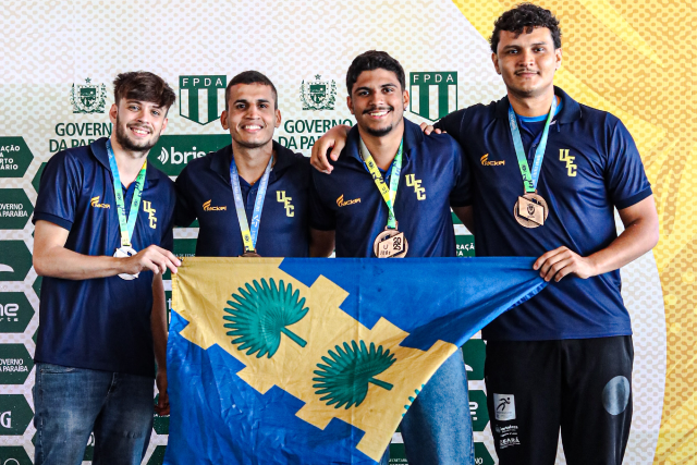Imagem: quatro estudantes homens da UFC seguram a bandeira com o brasão da universidade em frente ao banner nos jogos brasileiros universitários. eles vestem camisas na cor azul marinho e sorriem para a foto