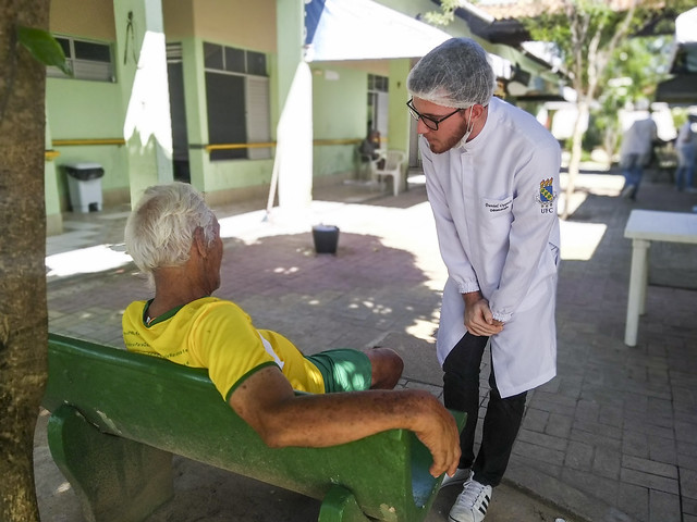 Na foto um estudante de jaleco branco conversa com um idoso de camisa amarela. Fim da descrição.
