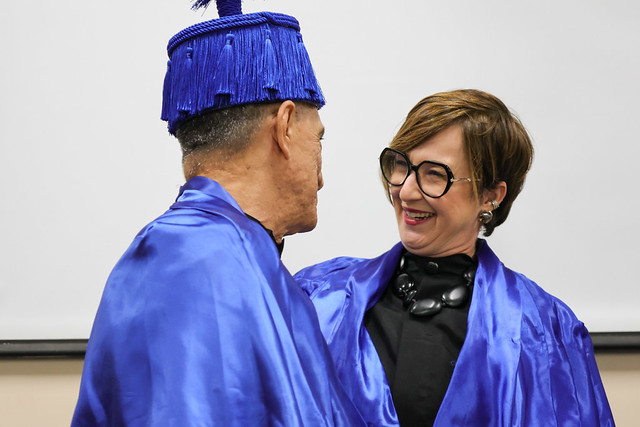 A professora Denise Parra cumprimenta o homenageado: "Flávio sistematizou um saber que rompe com modelos excludentes", pontuou a docente em seu discurso (Foto: Viktor Braga/UFC) Imagem: Um homem idoso e uma mulher se cumprimentam sorrindo. Ambos vestem becas azuis; ele usa um barrete azul, e ela, óculos grandes e colar preto.