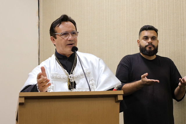 No discurso que encerrou a noite, o reitor Custódio Almeida recordou momentos importantes da trajetória profissional de Flávio Sampaio, ressaltando a natureza diversa e fundamental de suas contribuições (Foto: Viktor Braga/UFC) Imagem: Um homem veste beca branca e fala em um púlpito, gesticulando com as mãos. Ao lado dele, um intérprete de Libras, de camiseta preta, traduz o discurso.