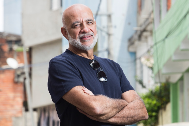 Um homem pardo careca de barba e camisa polo azul marinho posa para foto de braços cruzados