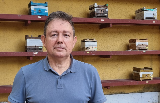Foto de um homem branco com camisa azul gola polo. Atrás dele, há prateleiras de madeira