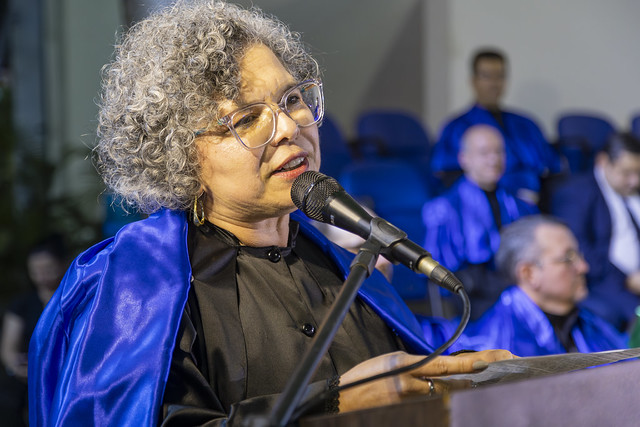 Imagem: uma mulher de cabelos cacheados e grisalhos, vestida de veste talar, fala em um púlpito