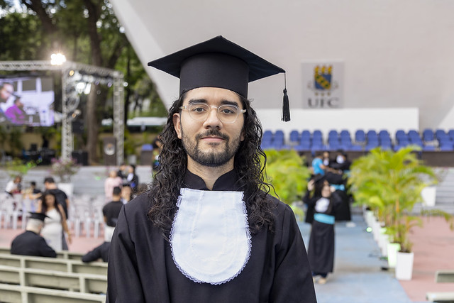 Imagem: um jovem branco de cabelos longos e vestido de beca posa para foto. Ao fundo, imagem do auditório da Concha Acústica