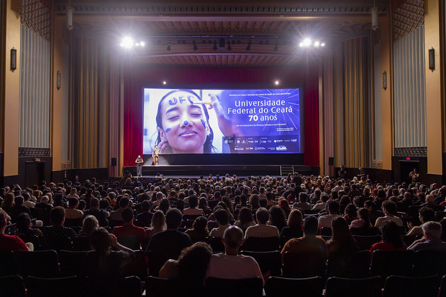 Foto de uma sala de cinema lotada. Ao fundo, na tela, a imagem de uma jovem com rosto pintado. Ao lado dela, sob fundo azul, está escrito Universidade Federal do Ceará 70 anos