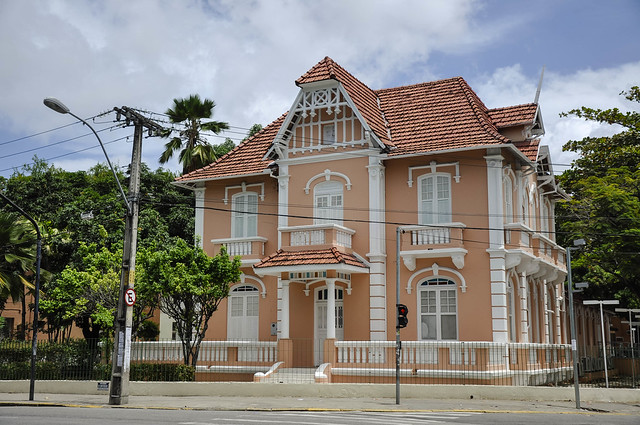 Imagem: Foto da fachada de um sobrado rosa com detalhes branco. Ao lado dele, há uma árvore e um poste de iluminação pública